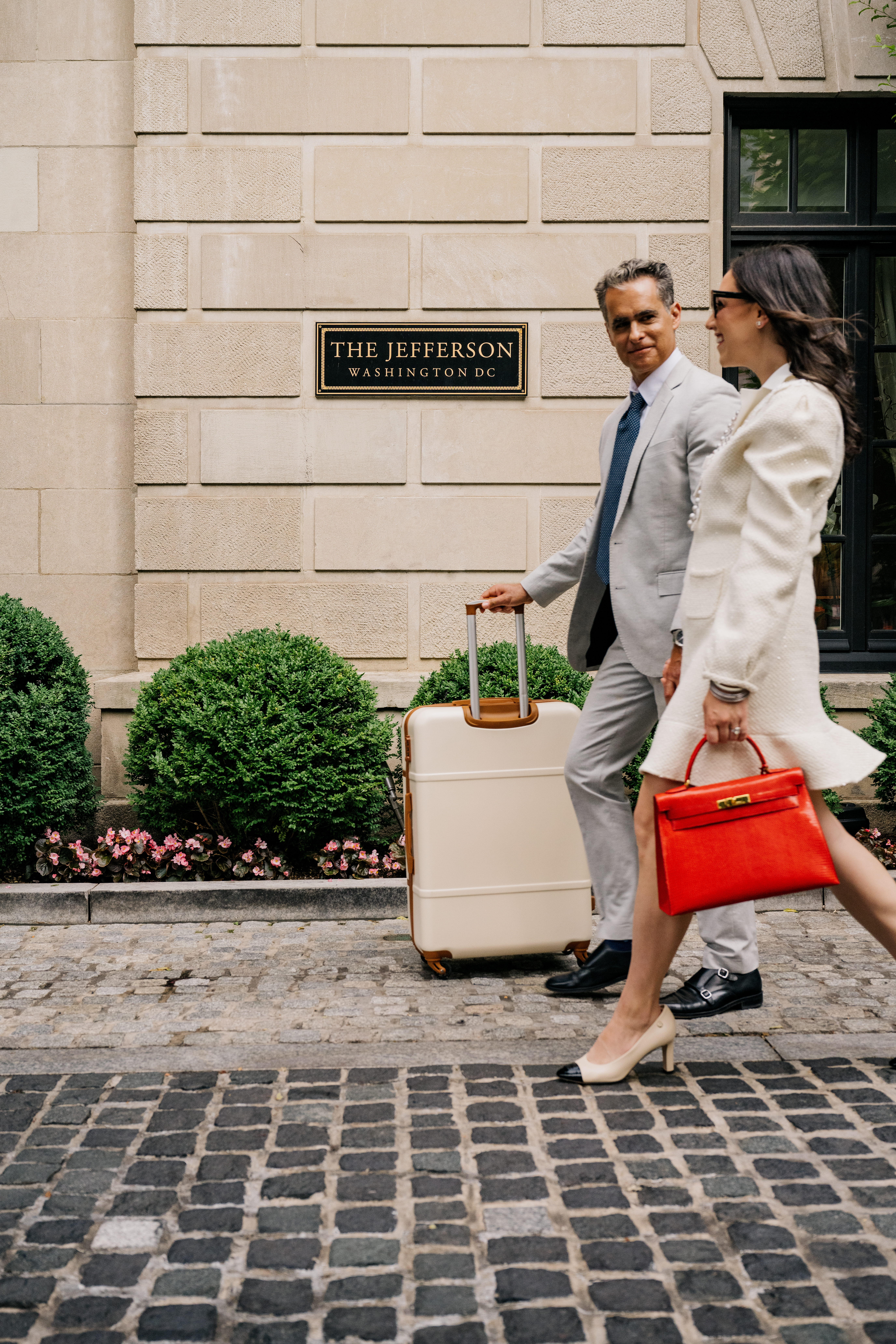 Couple walking outside hotel with suitcase and red handbag