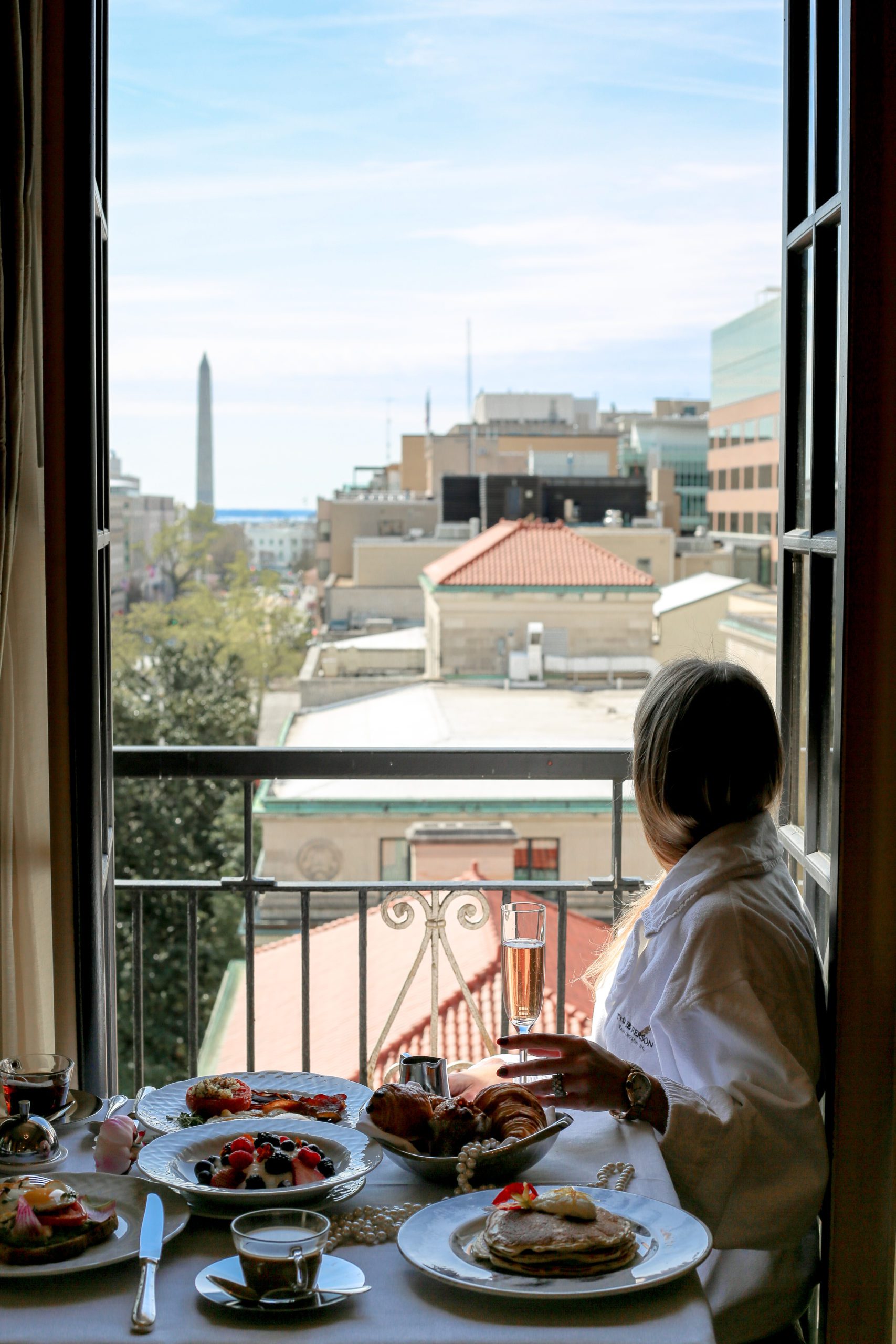 Person gazing out a sunlit window with a view of city rooftops and a distant monument.